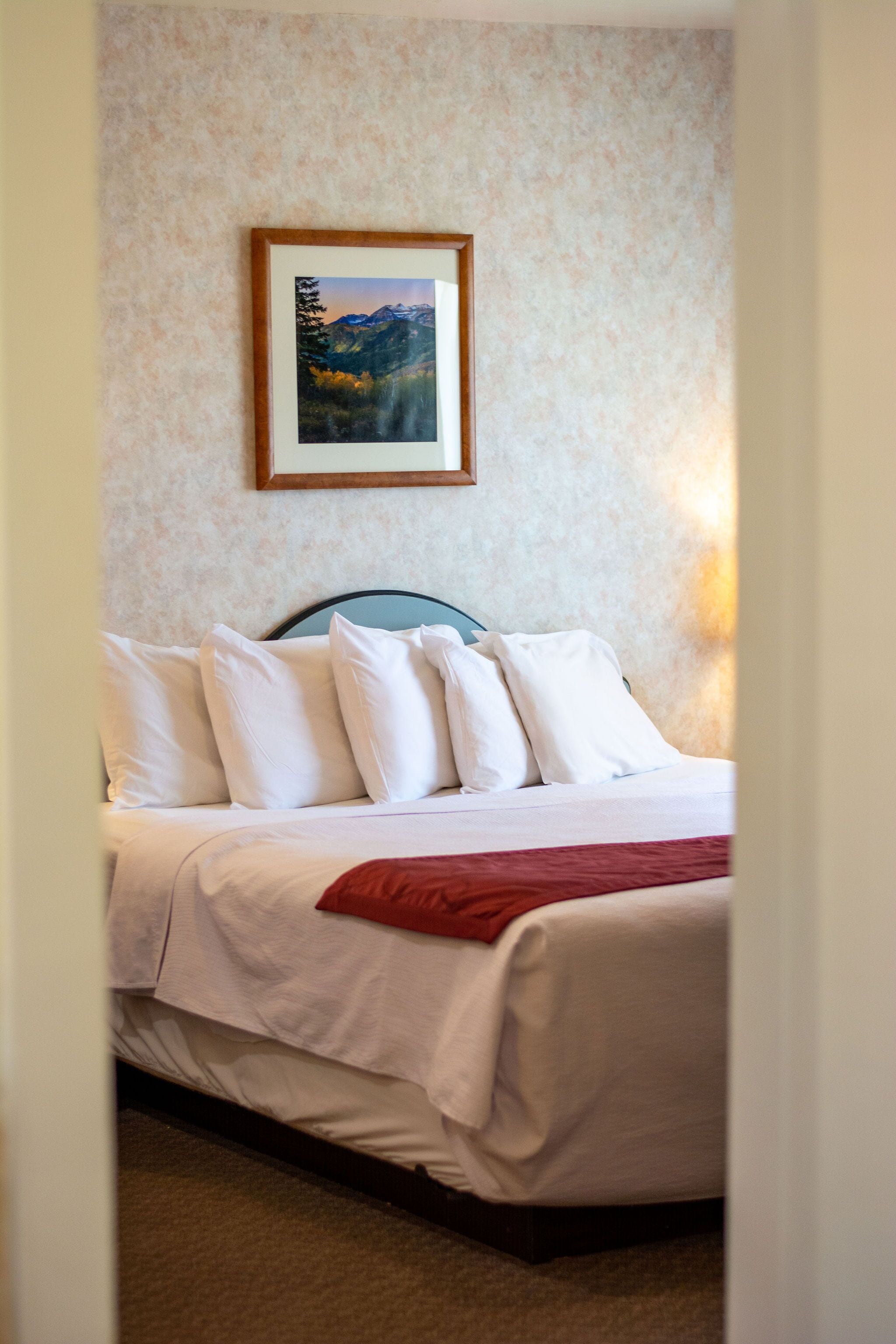 Un letto rifatto con lenzuola bianche e una coperta rossa, un quadro appeso alla parete e un pavimento con moquette.
