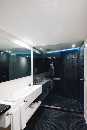 Un baño moderno con un espejo grande, tocador blanco y zona de ducha con baldosas oscuras.