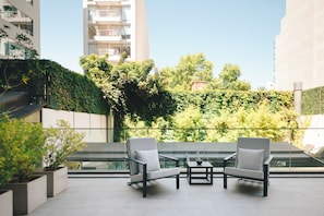 Un espacio exterior moderno con dos sillas y una mesa, rodeado de follaje y vistas a una piscina.