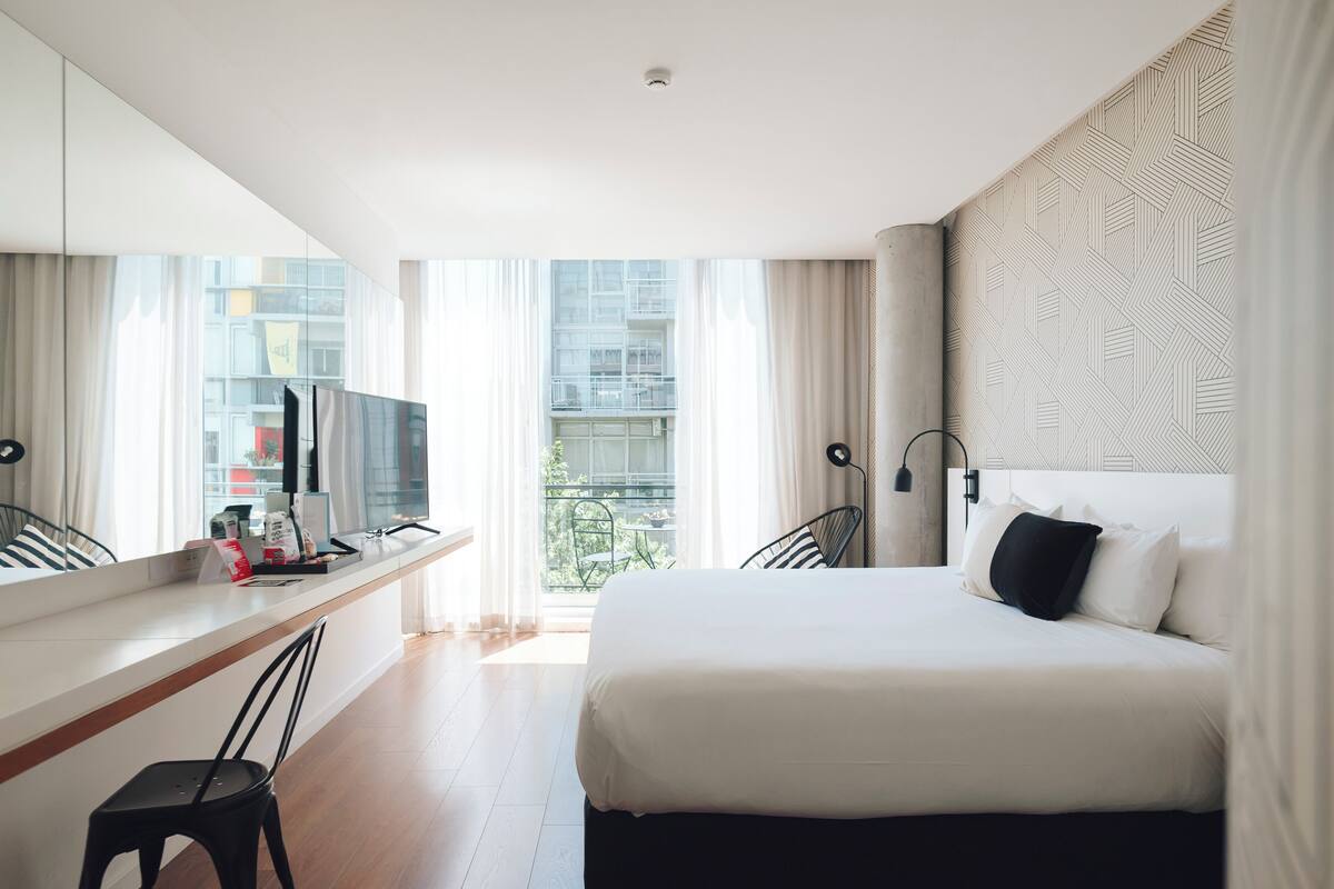 A modern hotel room with a large bed, a desk with a chair, a TV, and a balcony view.