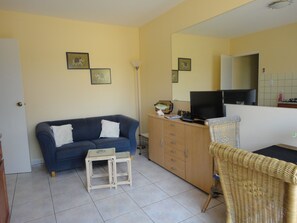 A living room with a blue sofa, a small white table, a wicker chair, a TV on a wooden cabinet, and a mirror on the wall. - Alta Montaña Apartments (Noord)