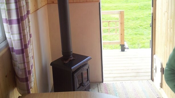 A room with a wood-burning stove, a wooden table, and a view of a grassy field through an open door. - Shepherds Lodge (Worcester)