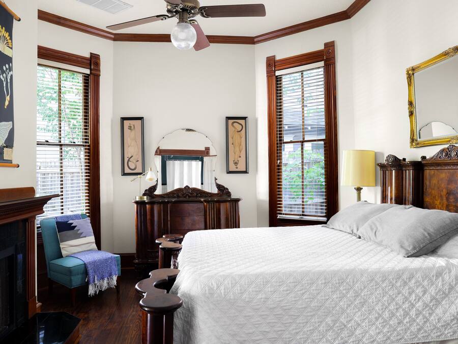 Een slaapkamer met een groot bed, een stoel met een plaid, een dressoir, een spiegel en een open haard.
