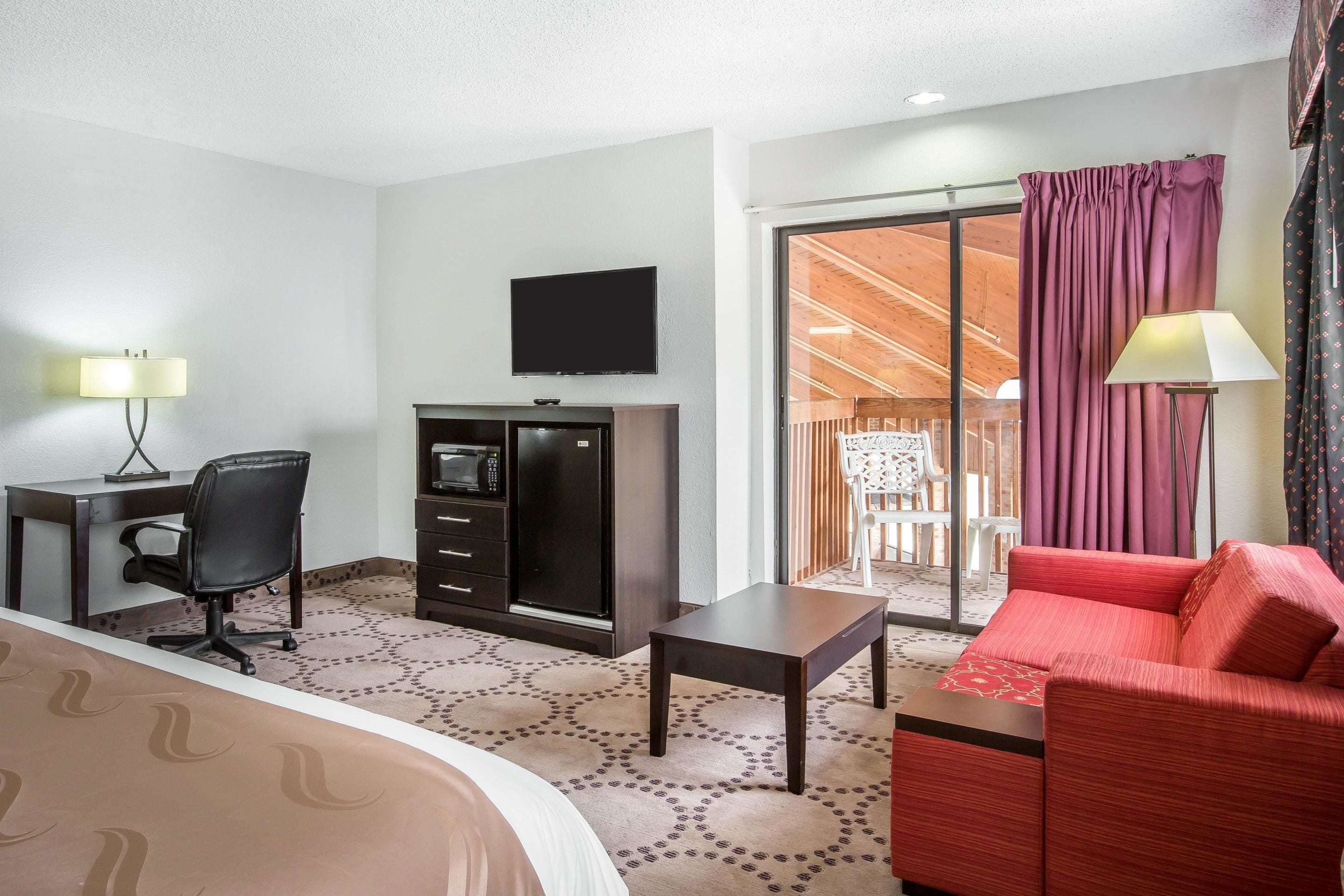 A hotel room with a bed, a desk with a chair, a TV, a small table, a red sofa, and a balcony with curtains.