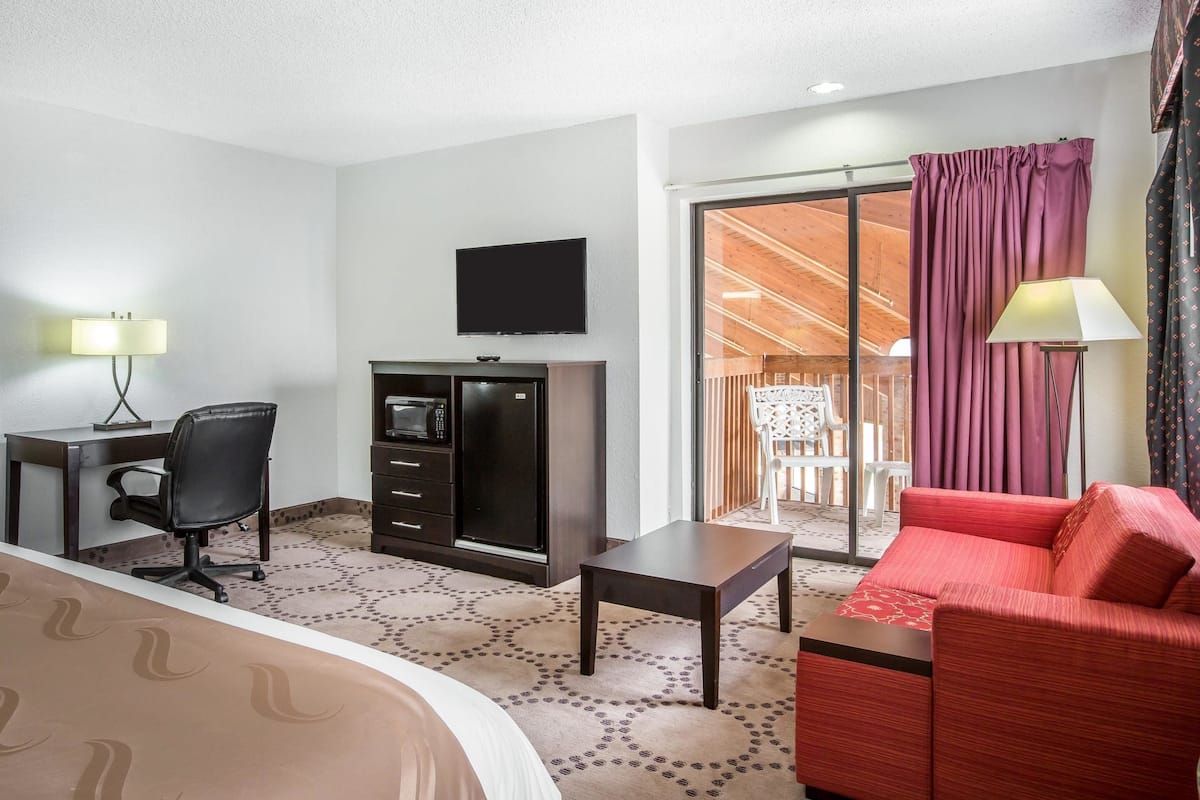 A hotel room with a bed, a desk with a chair, a TV, a small table, a red sofa, and a balcony with curtains.
