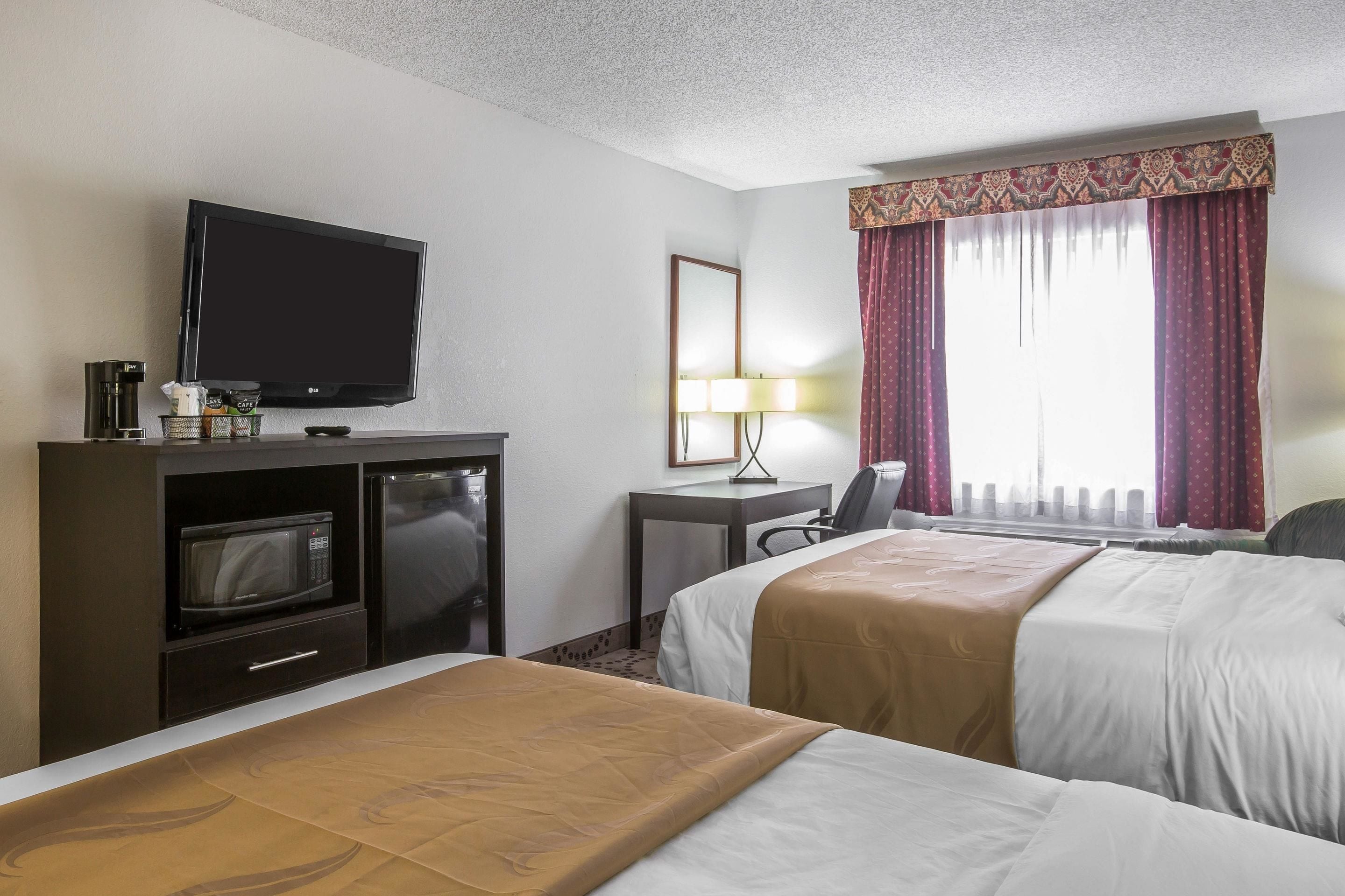 A hotel room with two beds, a TV, a desk with a lamp, and a window with curtains.