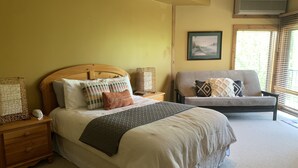 A bedroom with a wooden bed, multiple pillows, a bedside table with a lamp, a chair, and a window with blinds. - Longhollow Point Inn Studio 1105 (Galena)