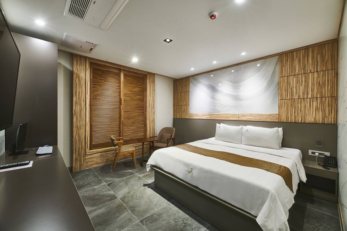 A modern hotel room with a large bed, a desk with a computer, a chair, and a wooden wardrobe.