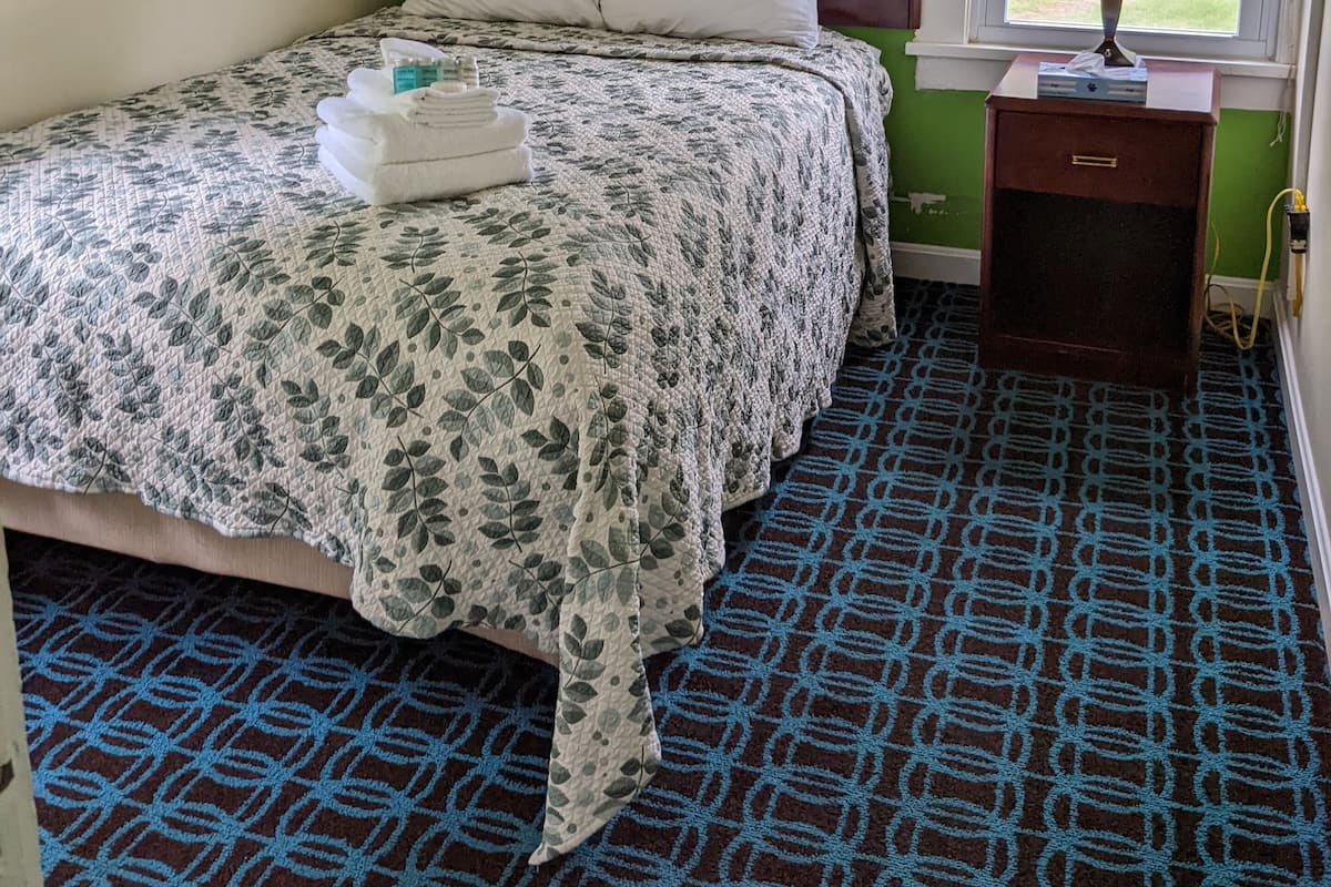 Uma cama de solteiro com um edredom e almofadas estampadas, uma mesinha de cabeceira com abajur e uma janela com vista para o exterior.