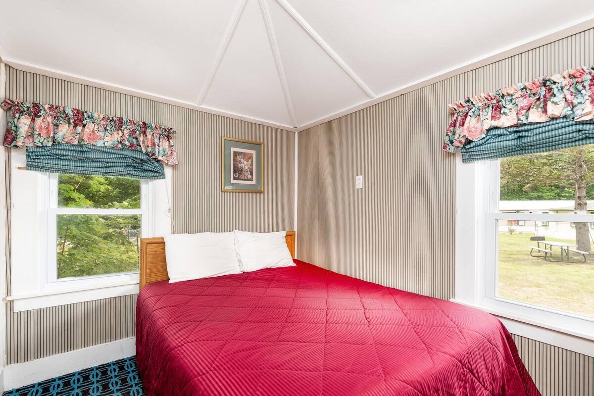 Quarto com colcha vermelha, papel de parede listrado e baldrame floral nas janelas.