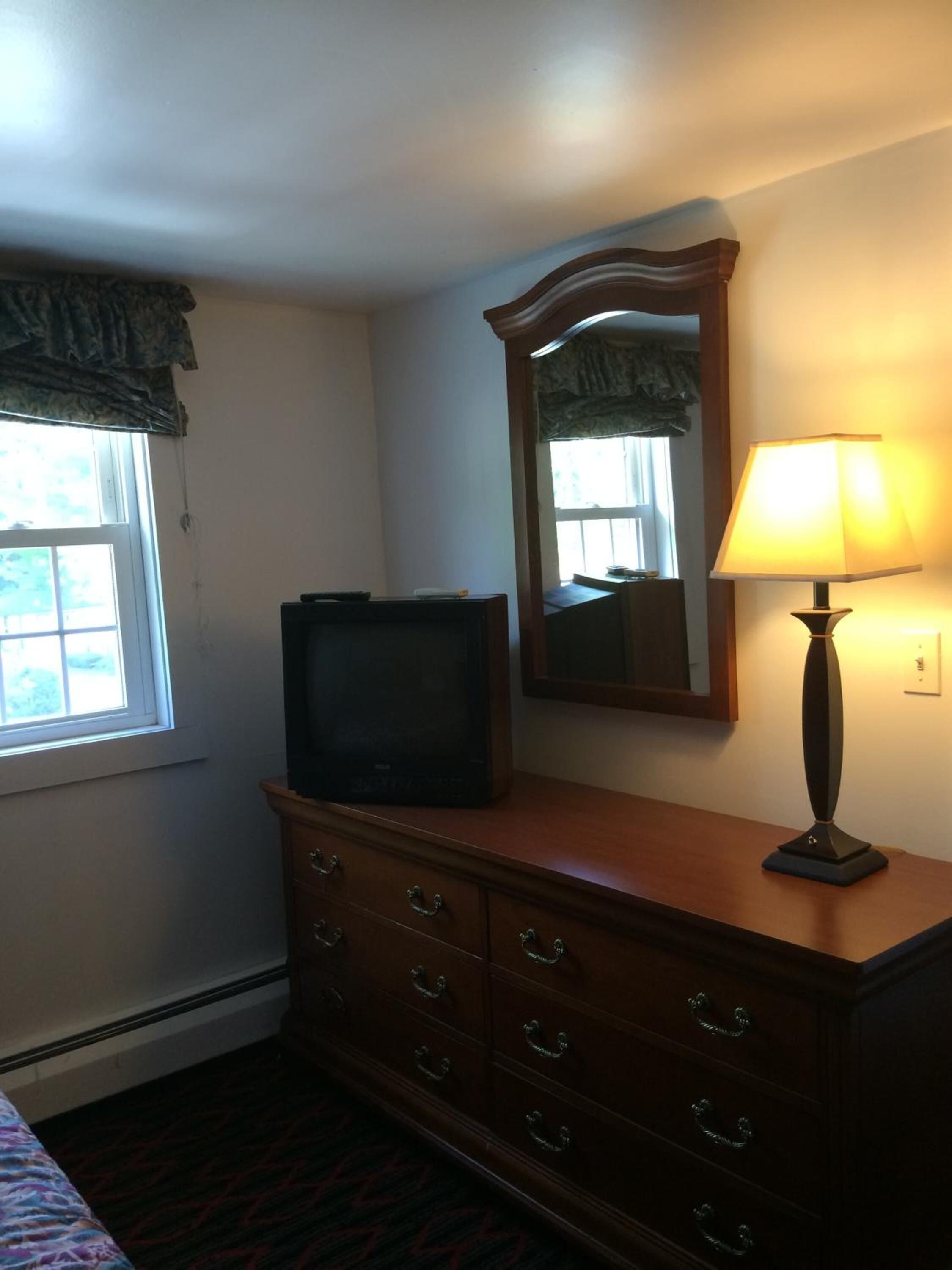 A room with a wooden dresser, a television on top, a mirror, and a lamp.
