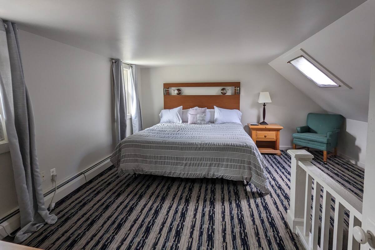 Quarto de hotel com cama, mesa de cabeceira, luminária, cadeira e claraboia.
