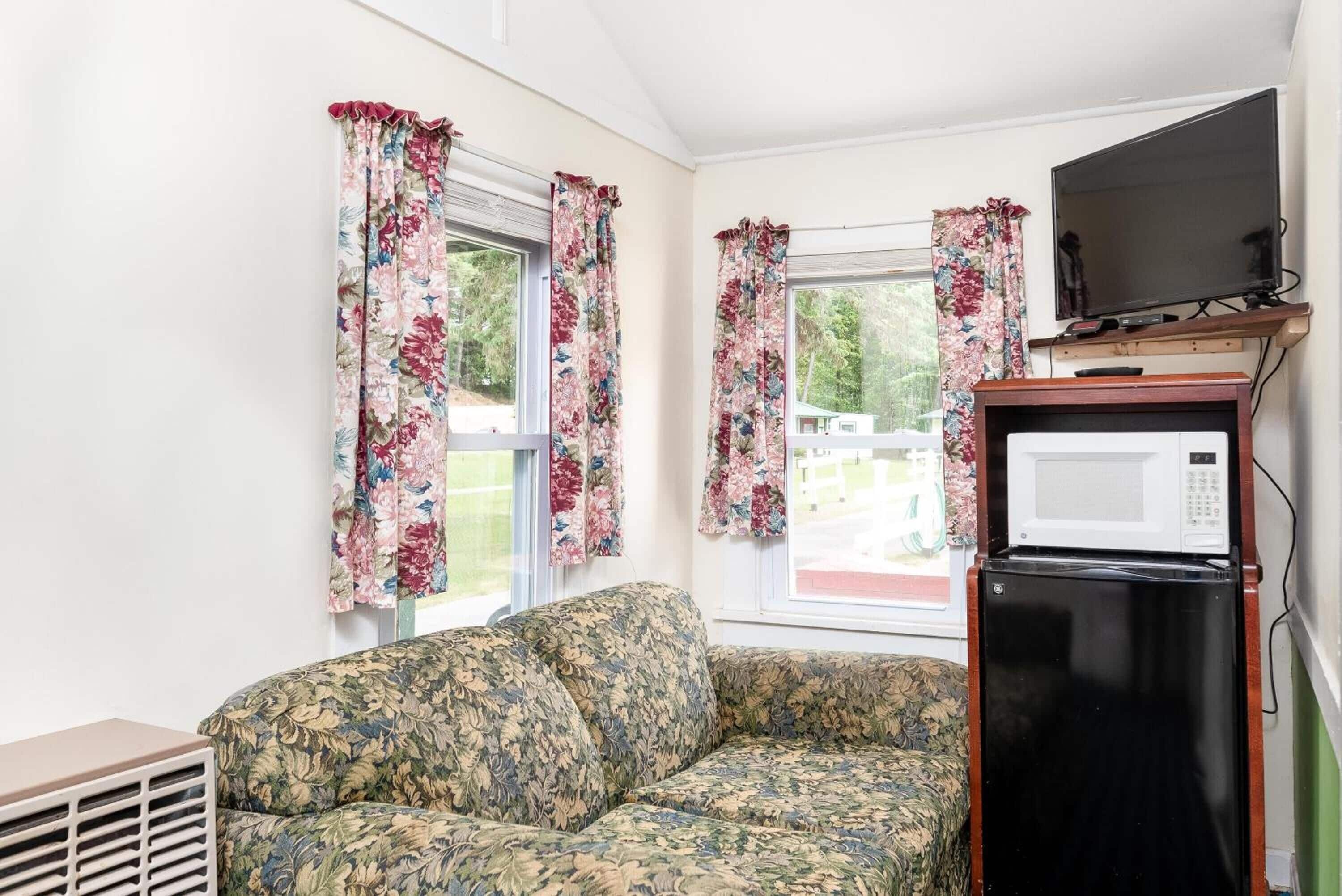 A room with a floral-patterned sofa, a black refrigerator, a television on a wooden stand, and windows with floral curtains.