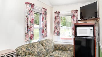 A room with a floral-patterned sofa, a black refrigerator, a television on a wooden stand, and windows with floral curtains.