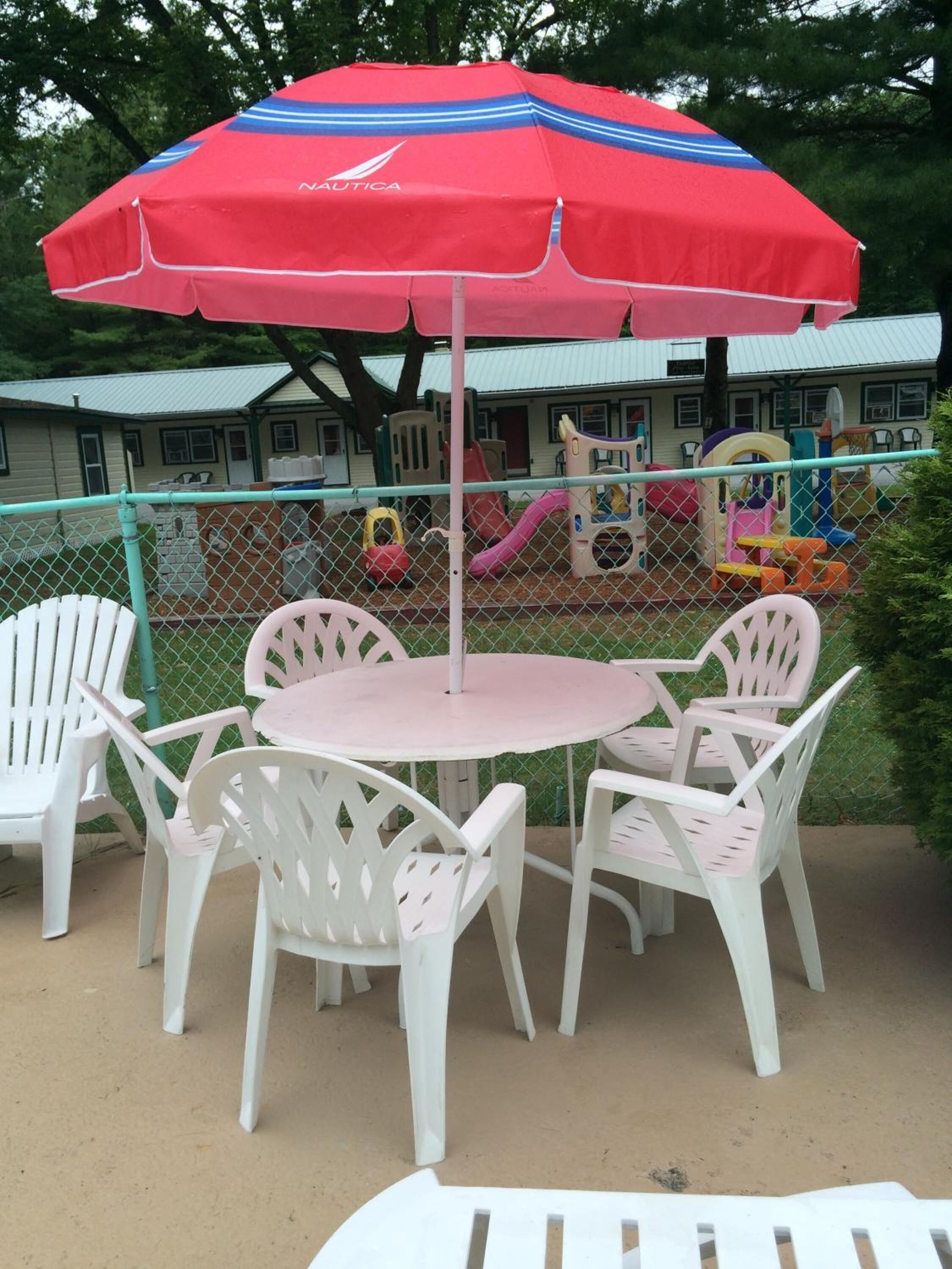 A patio area with a round table and four white chairs, a red umbrella, and a playground in the background.