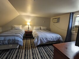 Una habitación pequeña y bien iluminada con dos camas, una mesita de noche con lámpara, alfombra estampada y un equipo de aire acondicionado en la pared.