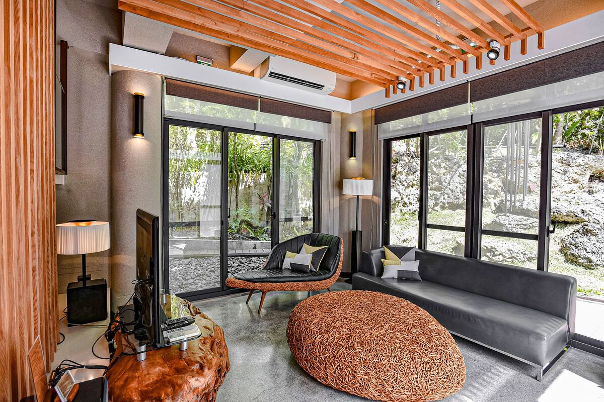A modern living room with large windows, a sofa, a chair, and a unique woven ottoman.
