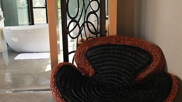Un interior moderno con una silla de mimbre y un reposapiés, una bañera de vidrio y un separador decorativo de metal negro.