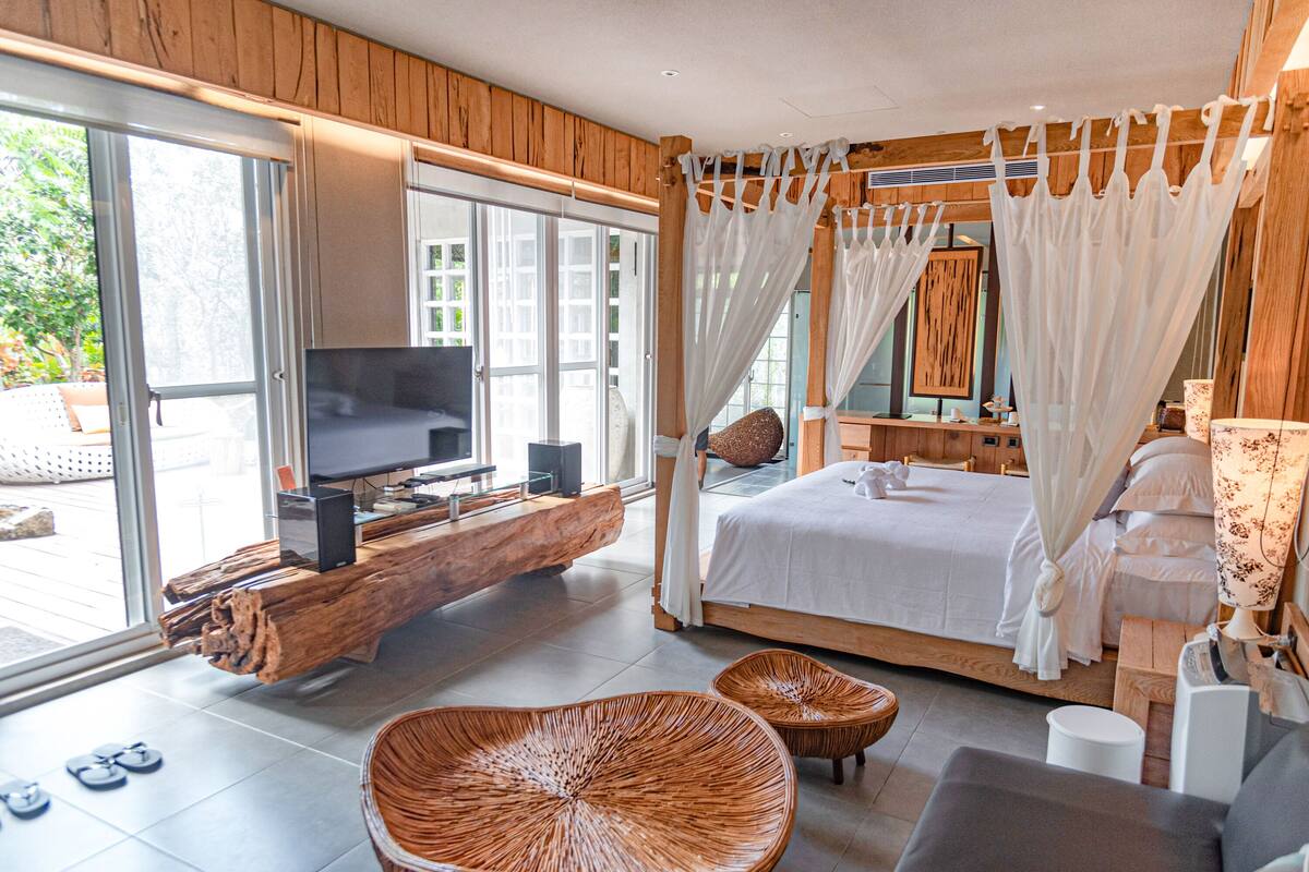 A modern bedroom with a large bed, a TV on a wooden shelf, wicker furniture, and a large window.