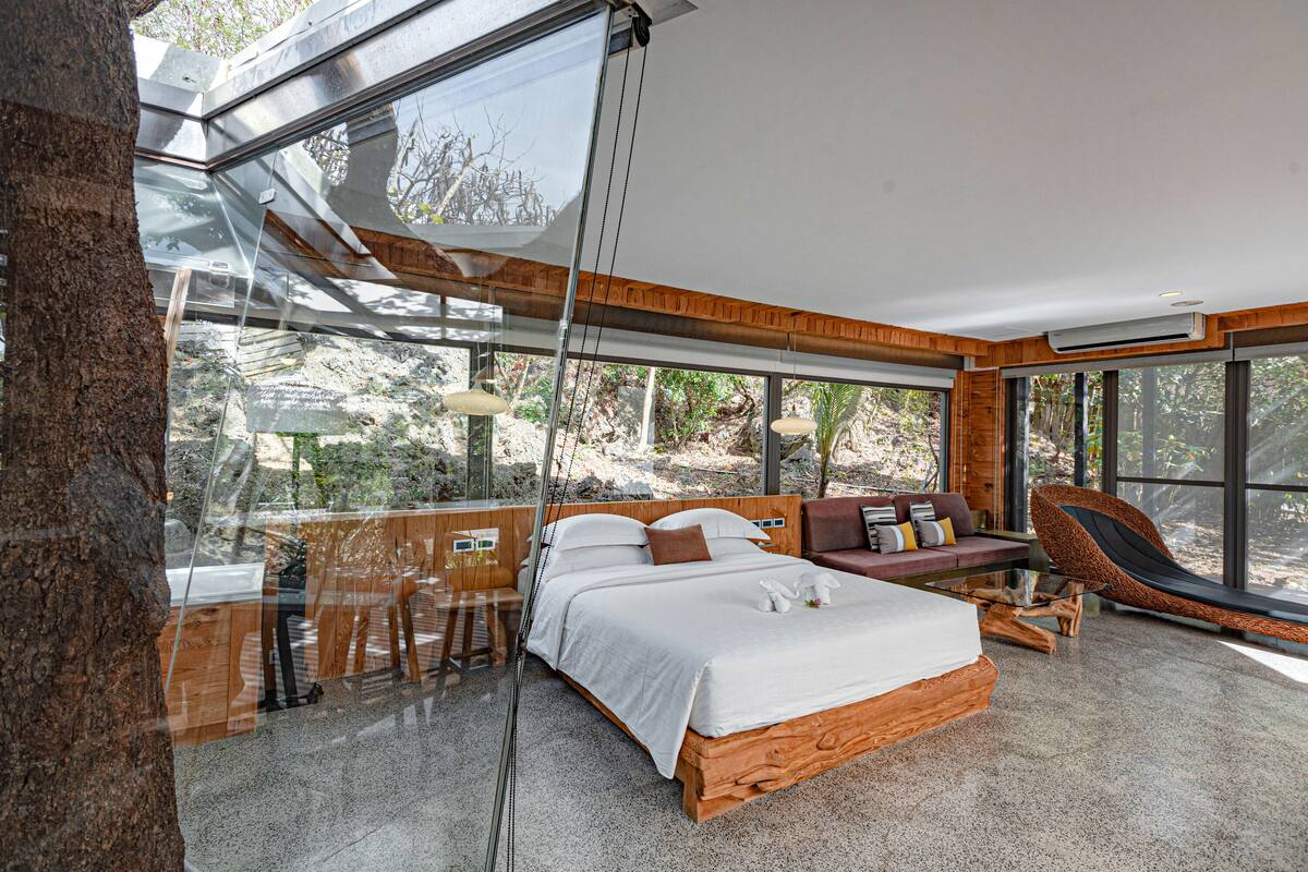 A modern bedroom with a large bed, wooden headboard, and a view of the outdoors through large windows.