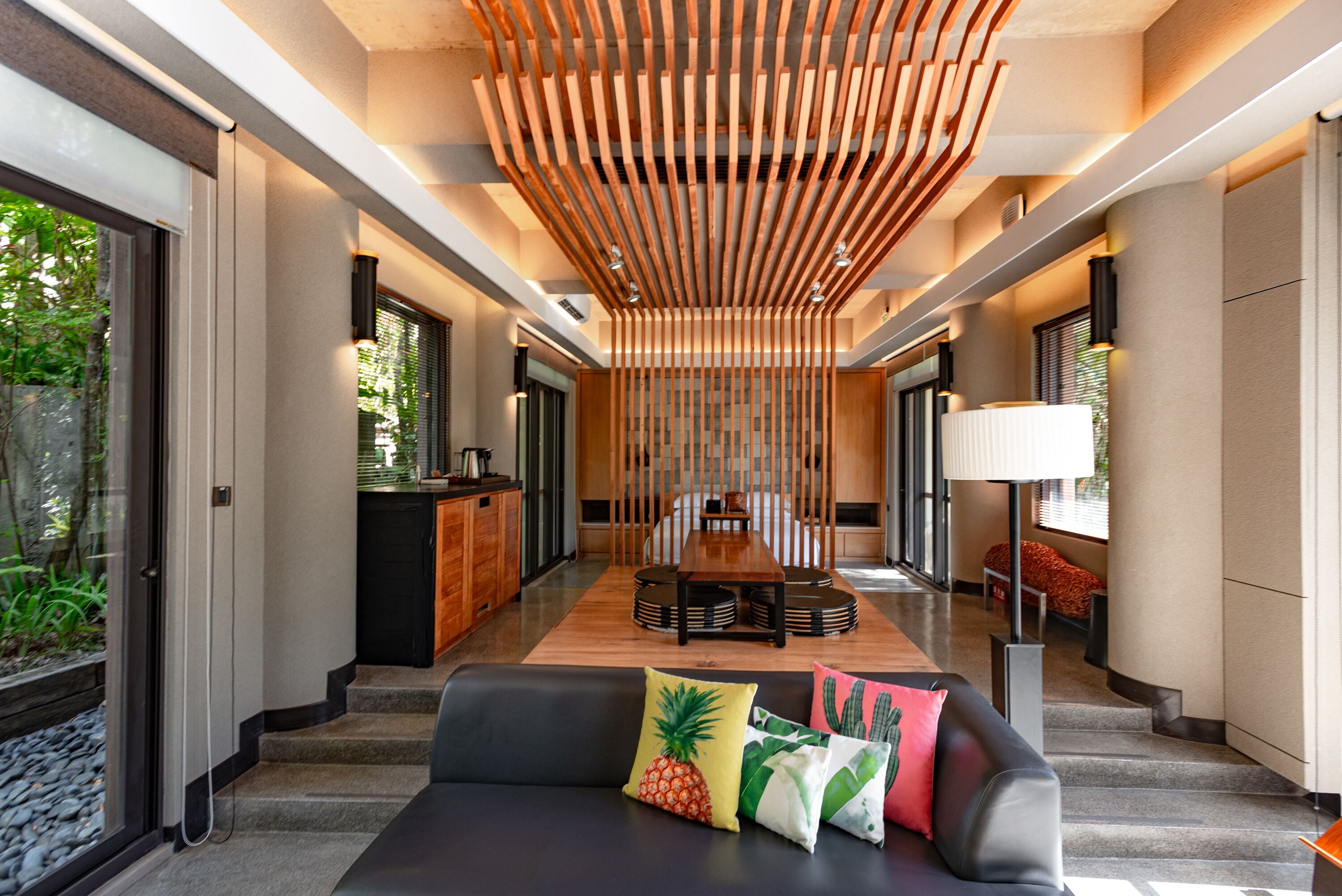 A modern interior with a central dining area, a sofa with colorful cushions, and a wooden ceiling structure.
