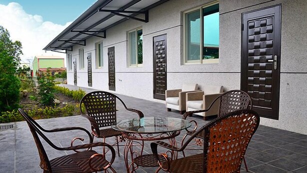 Un patio con mesa de vidrio y sillas de mimbre, un sofá, y un edificio con ventanas grandes y pared texturizada.