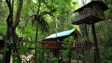 A treehouse with a metal balcony and a wooden deck, surrounded by dense forest.