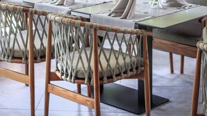 Un área de comedor al aire libre con sillas de mimbre y una mesa dispuesta para dos personas, con vistas a un follaje exuberante y un edificio moderno al fondo.