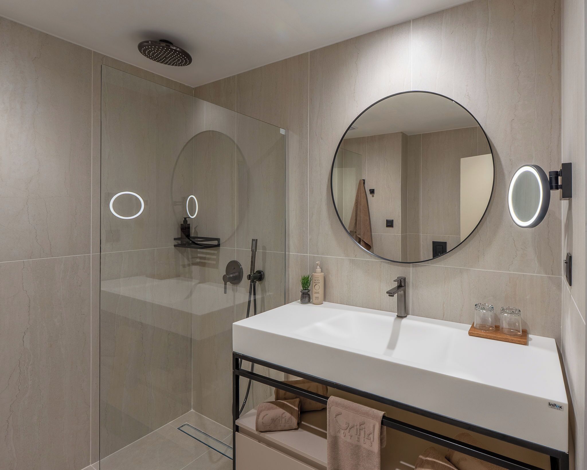 A modern bathroom with a glass shower enclosure, a round mirror, a white sink, and a towel rack.