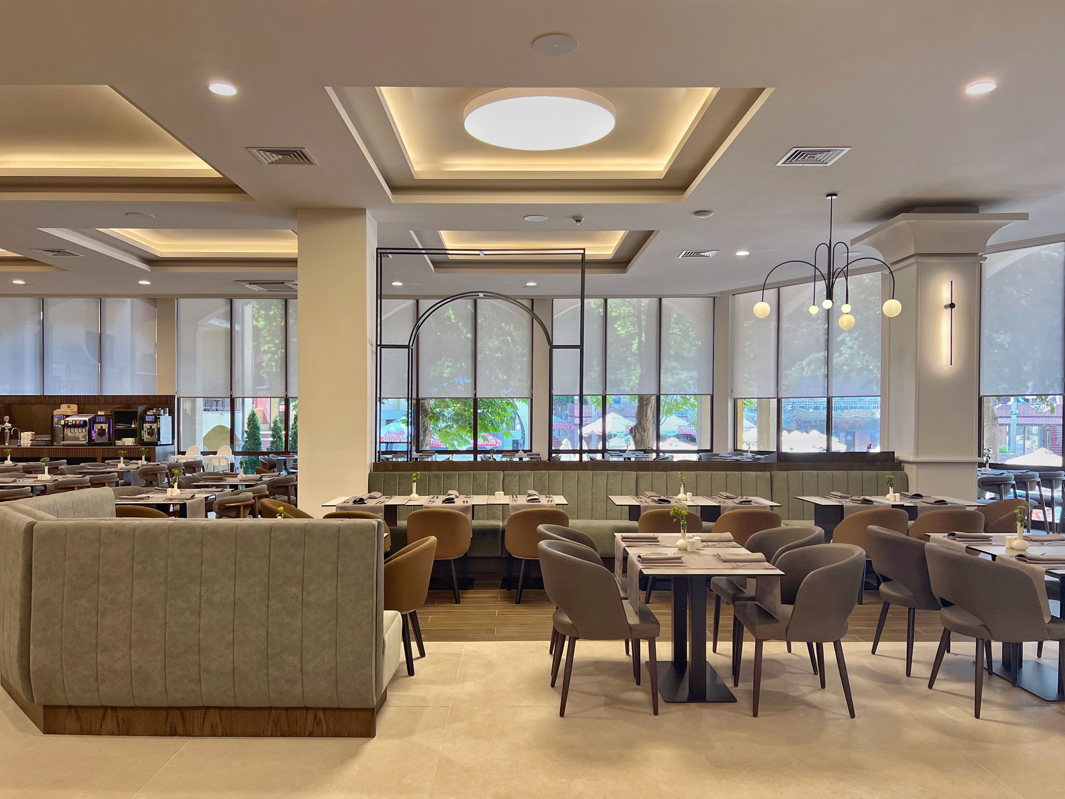 A modern restaurant with a central seating area, high ceilings with recessed lighting, and large windows offering a view of the outdoors.