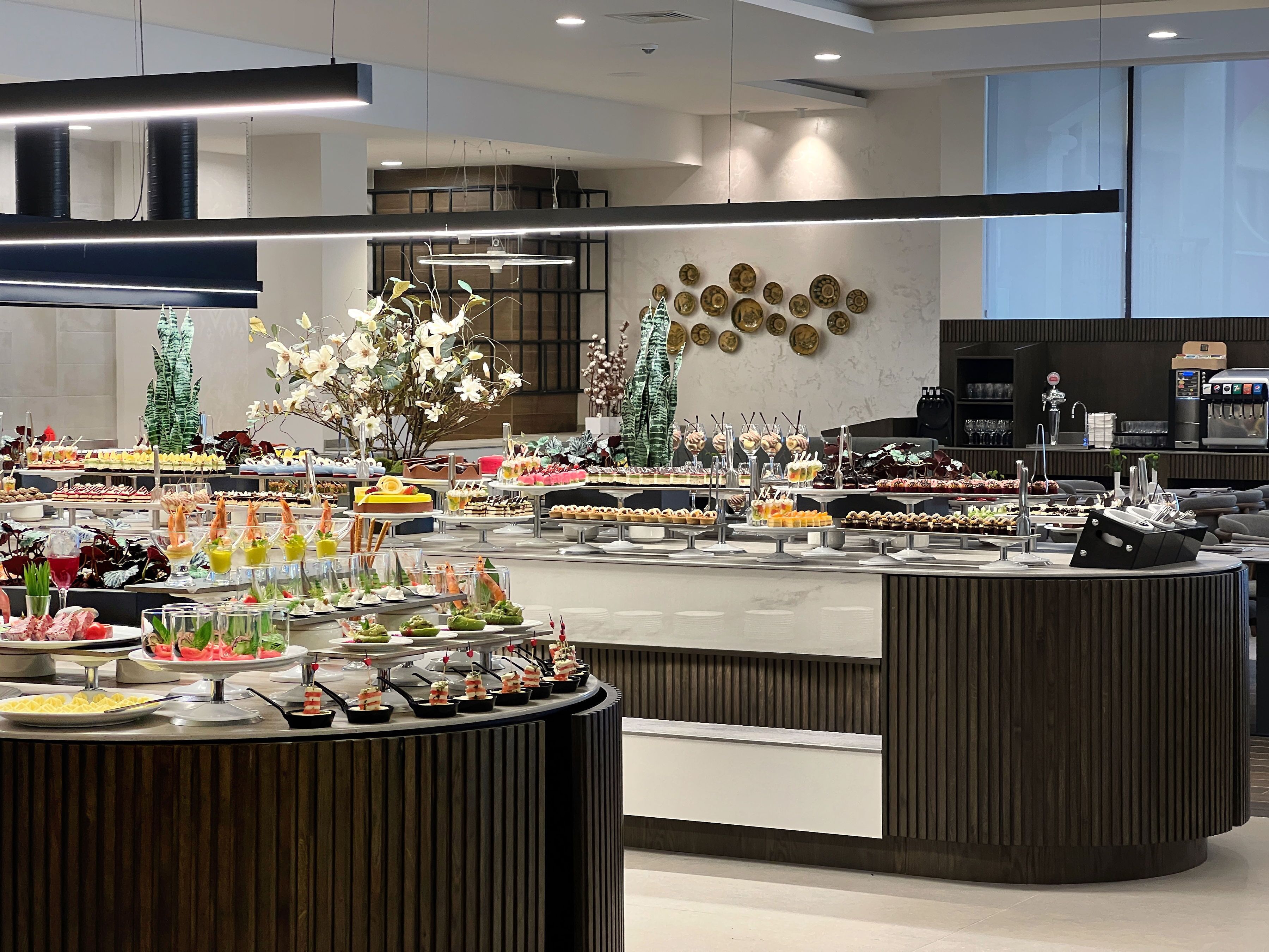 Un espace buffet avec différents stands de nourriture, une machine à café et des plantes décoratives.