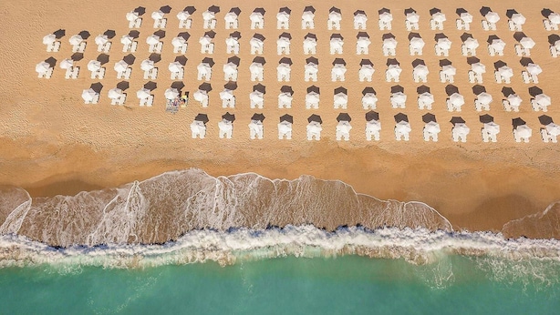 Vista aérea de una playa con hileras de sombrillas y sillas de playa blancas, una orilla arenosa y olas acercándose a la costa.