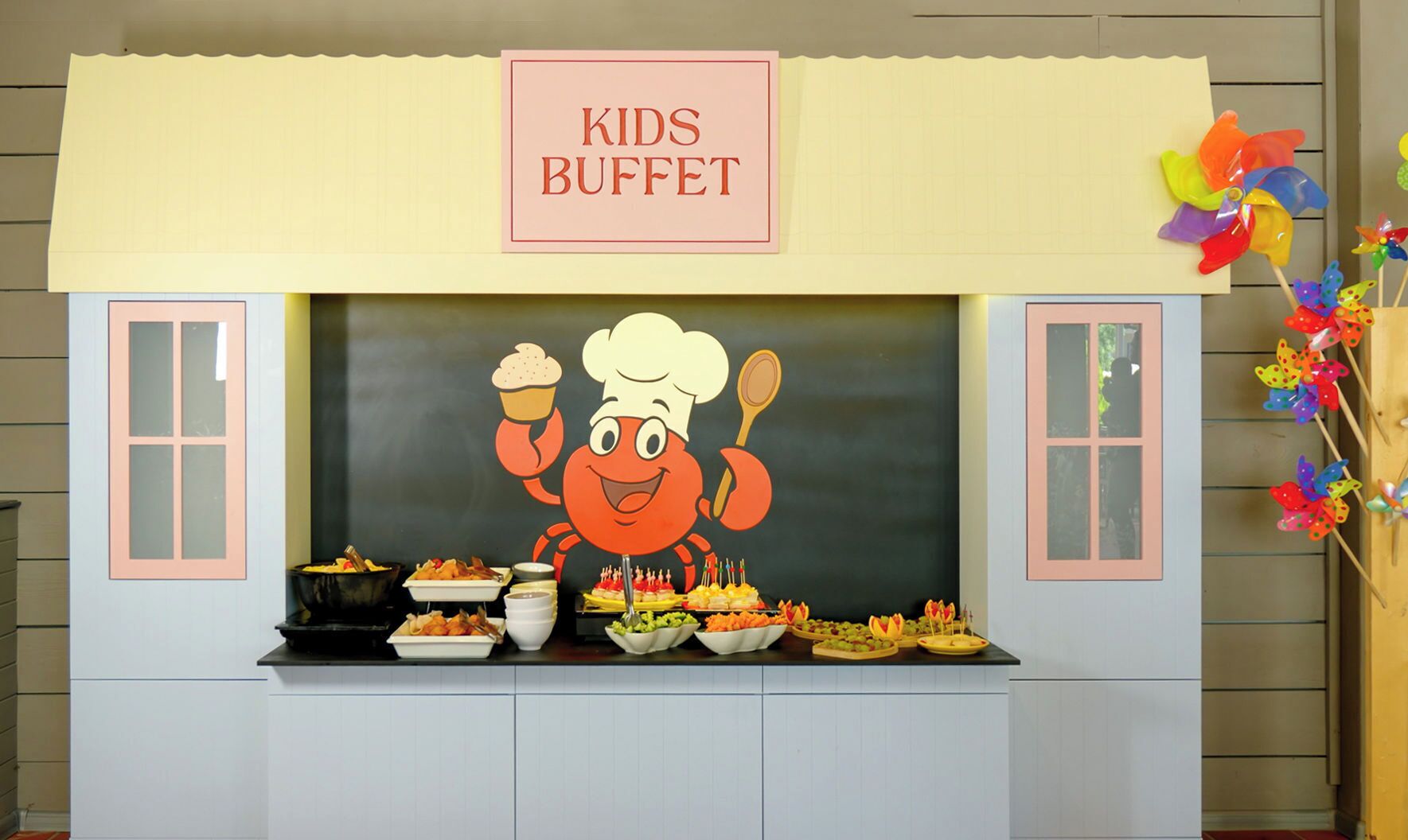 A kids' buffet area with a cartoon chef decoration, various food items, and pinwheel decorations.