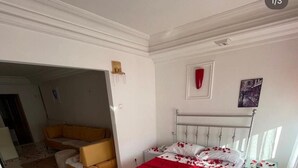 Un dormitorio con una cama cubierta de pétalos de rosa, una colcha roja y un cabecero blanco. Hay un banco de madera y una mesita con un jarrón de flores.