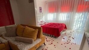 Un moderno salón con un sofá, una cama con ropa de cama roja y pétalos de rosa dispersos en el suelo.