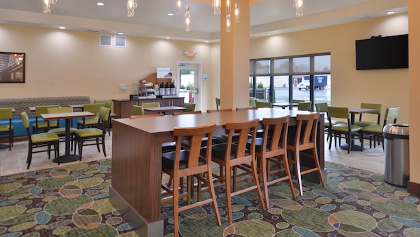 A spacious dining area with multiple tables and chairs, a central bar area, and a large window providing a view of the outside. - Holiday Inn Express & Suites Dearborn SW - Detroit Area by IHG (Dearborn)