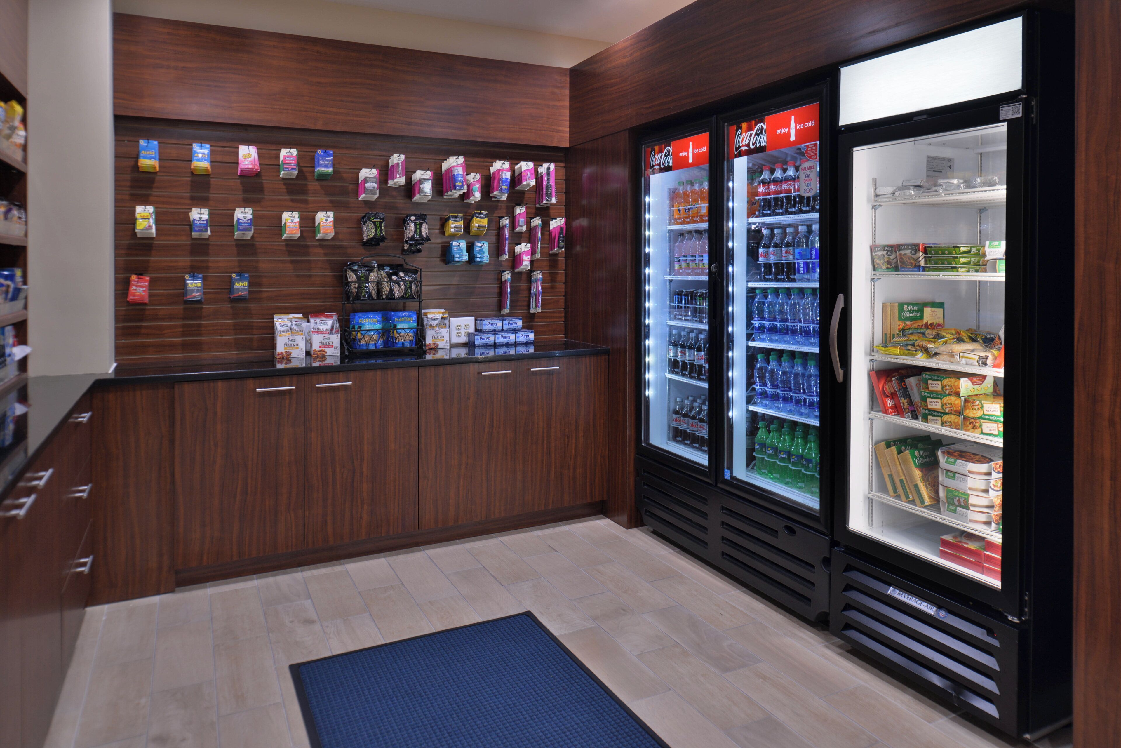 A hotel lobby with a wooden reception desk, a blue mat, and two beverage air conditioning units stocked with various drinks.