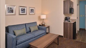 A hotel room with a blue sofa, a wooden coffee table, a lamp, a microwave, and a small kitchen area. - Holiday Inn Express & Suites Dearborn SW - Detroit Area by IHG (Dearborn)