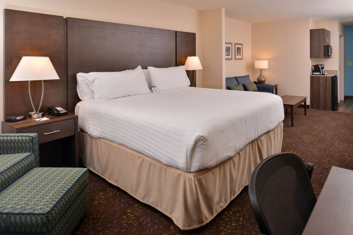 A hotel room with a large bed, two bedside tables with lamps, a desk with a chair, and a kitchenette area.