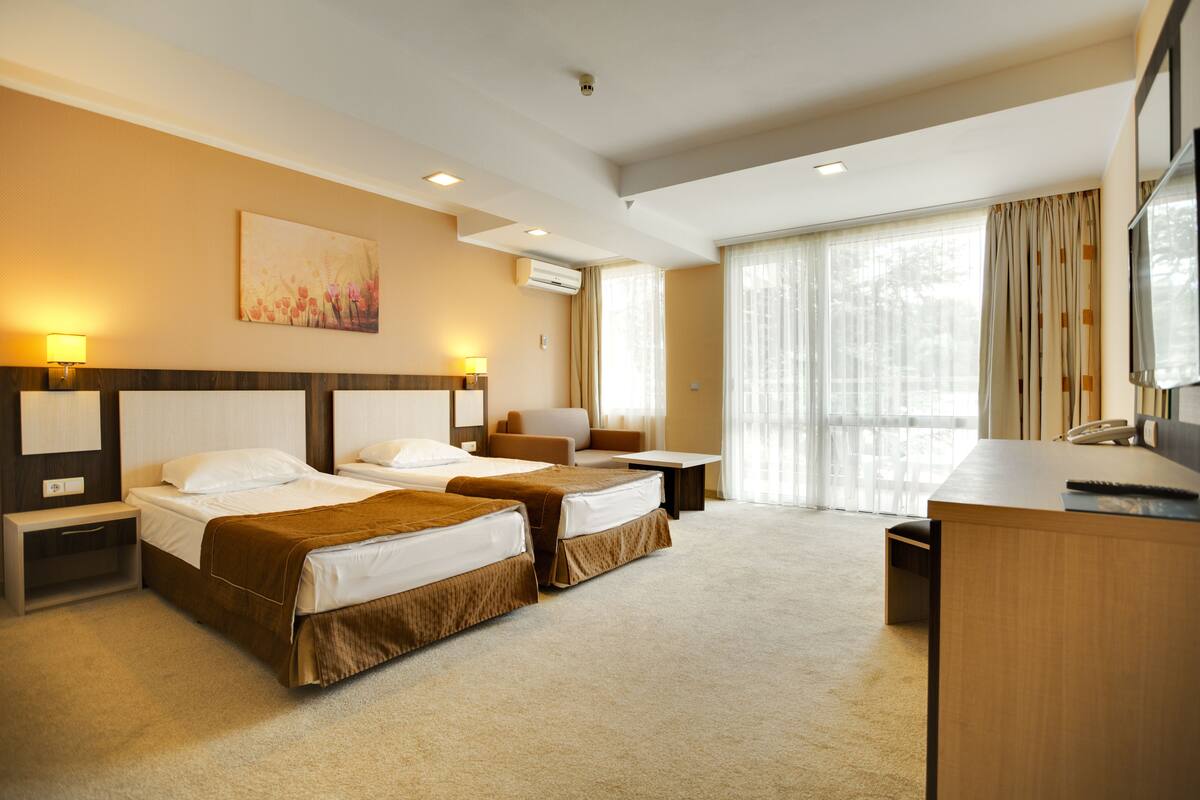 A hotel room with two beds, a desk, a chair, a TV, and a large window with curtains.