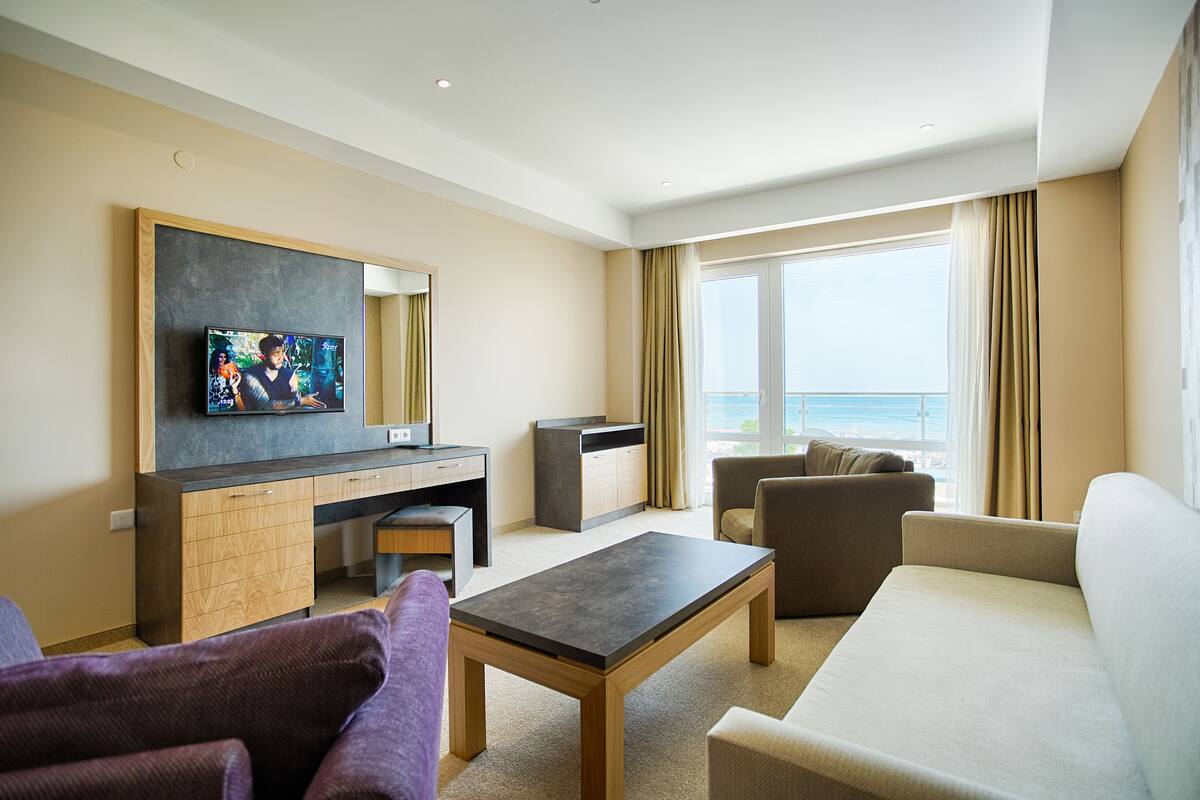 A modern hotel room with a large window, a flat-screen TV, a sofa, a coffee table, and a desk.