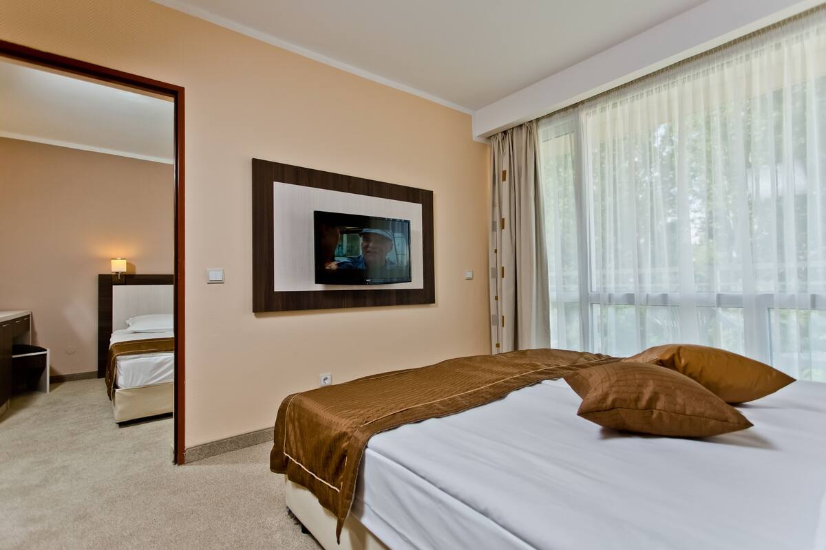 A hotel room with a bed, a television mounted on the wall, a bedside table, and a window with curtains.