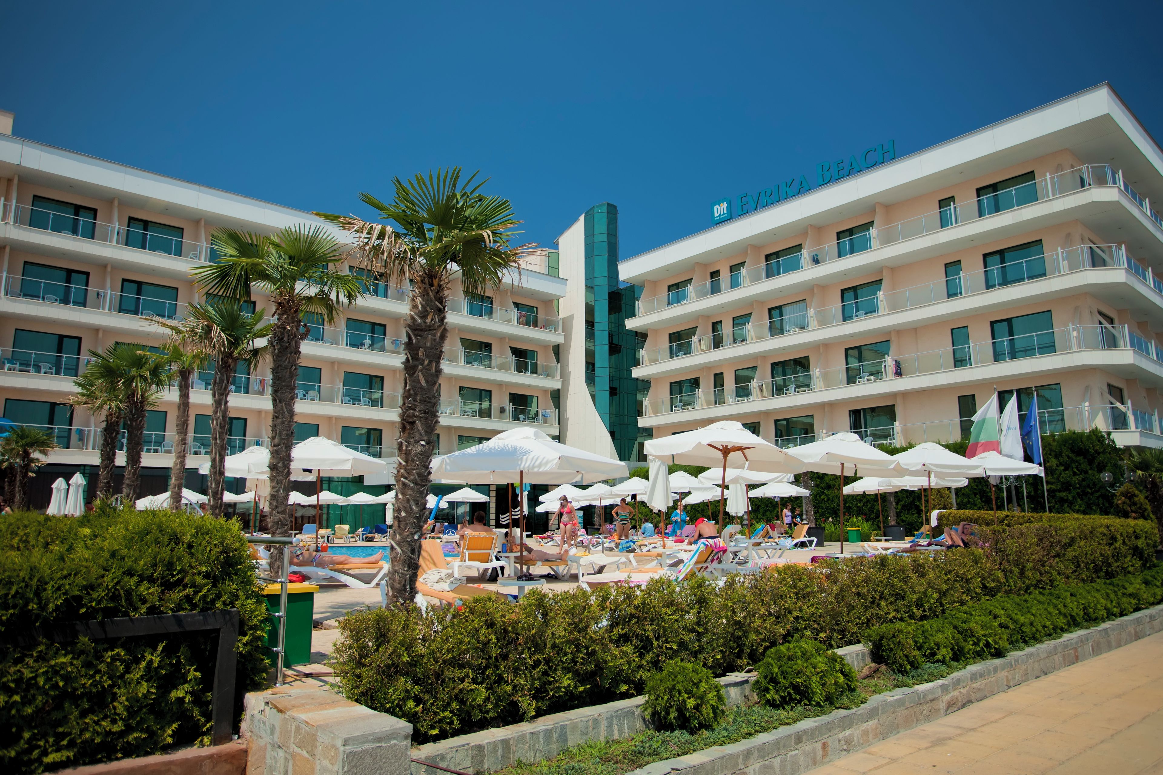 A hotel with multiple balconies, a pool area with sun loungers and umbrellas, and a clear blue sky.