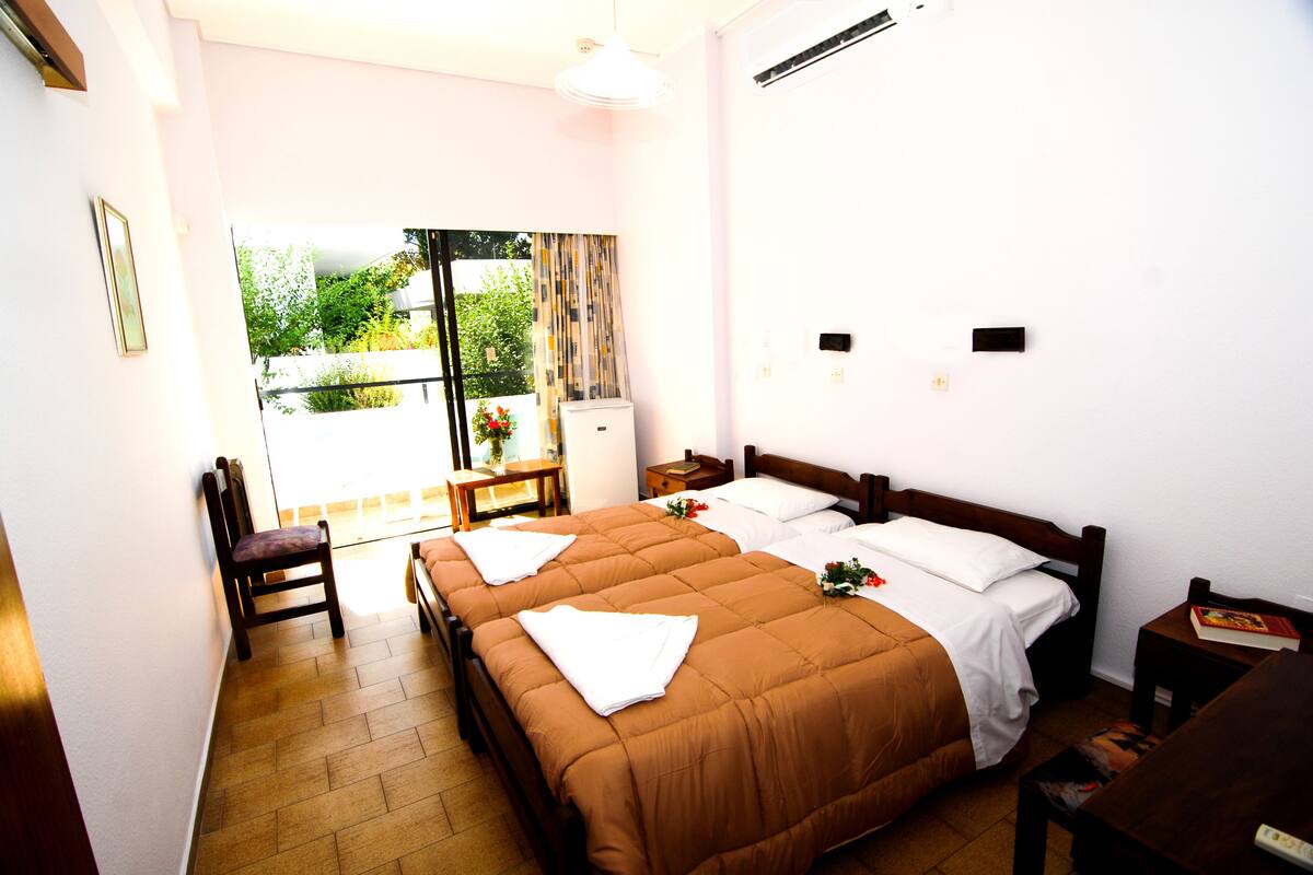 A hotel room with two beds, a wooden chair, a small table, and a view of greenery outside.