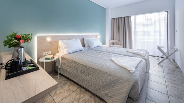 A hotel room with two beds, a bedside table with a lamp, a small side table, and a window with curtains. - Aegeon Hotel (Kos)