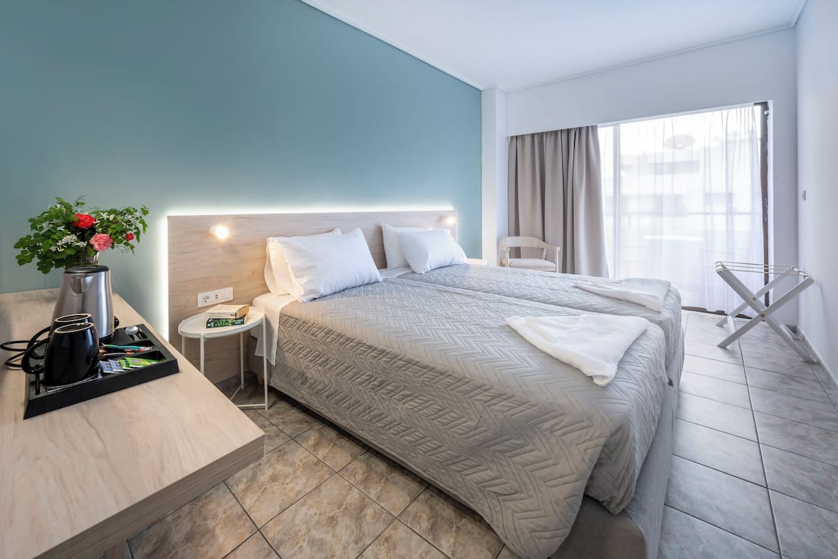 A hotel room with two beds, a bedside table with a lamp, a small side table, and a window with curtains.