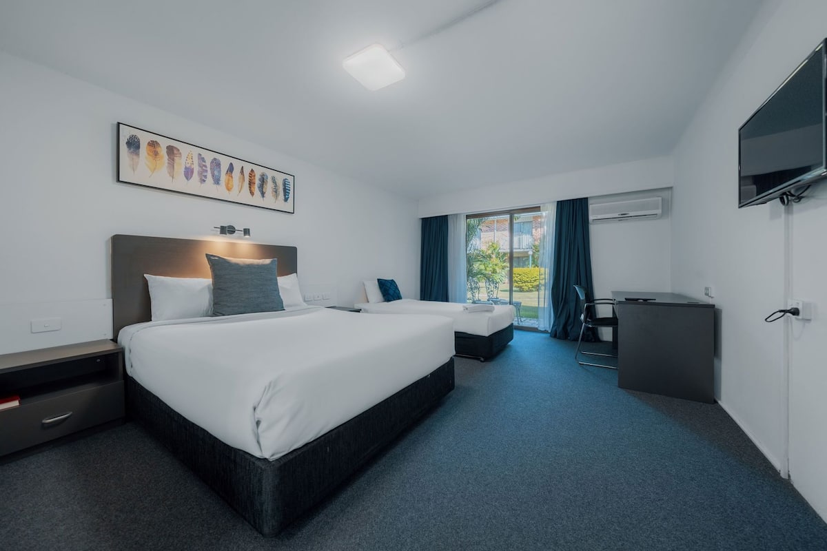 A hotel room with two beds, a desk, a TV, and a large window with curtains.