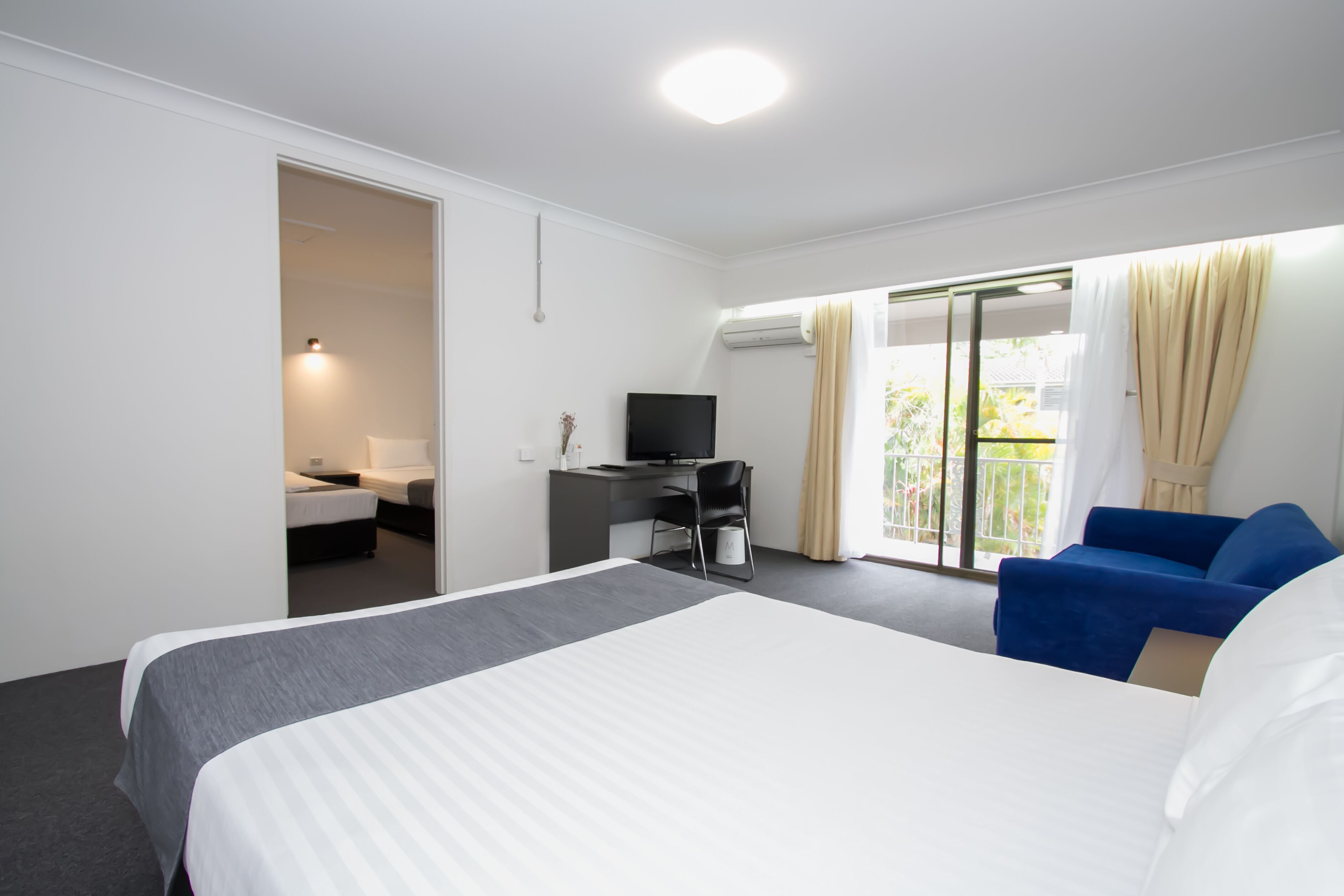 A hotel room with a large bed, a desk with a computer, a TV, and a balcony with curtains.