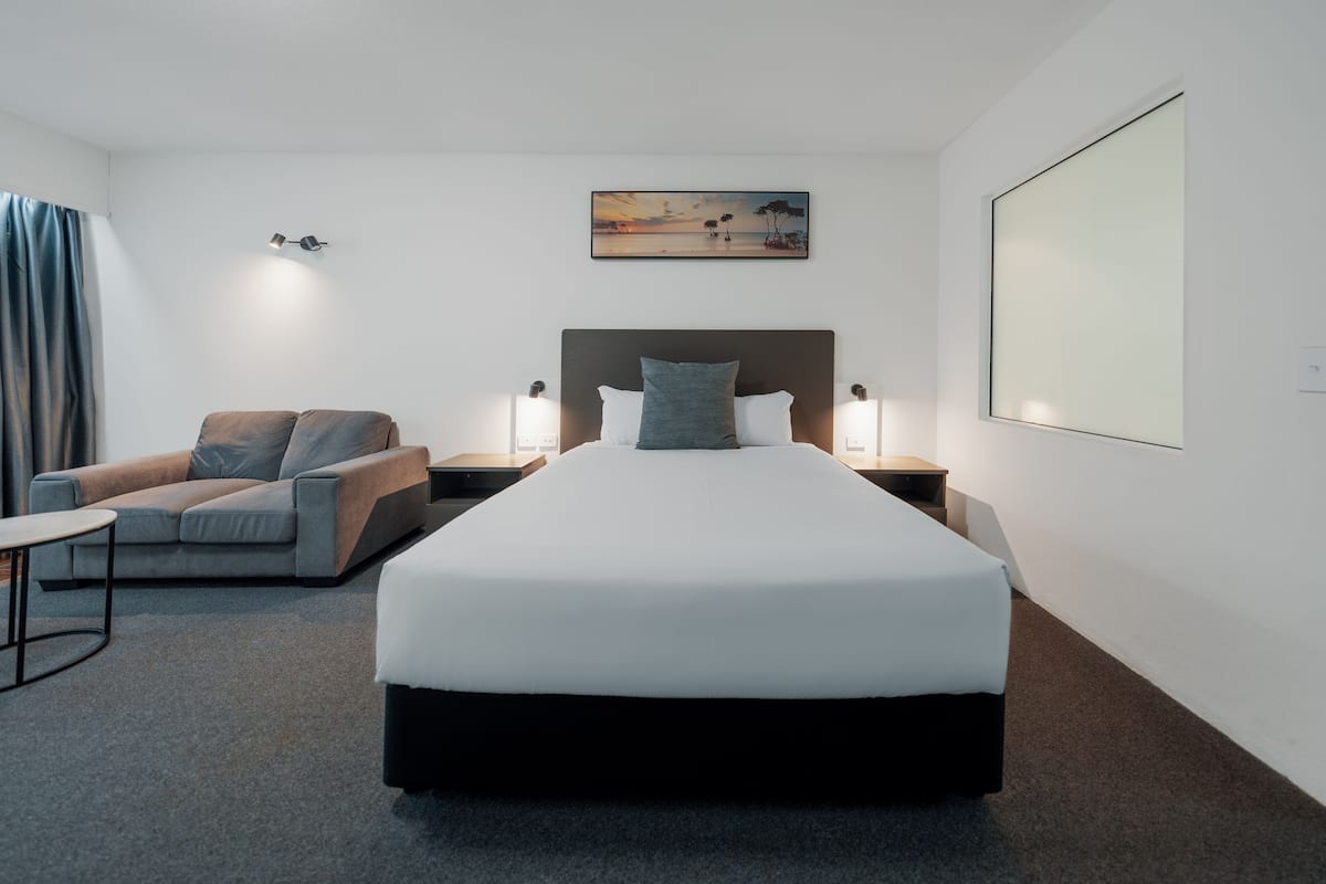 A hotel room with a large bed, a sofa, two bedside tables with lamps, a painting on the wall, and a large window.