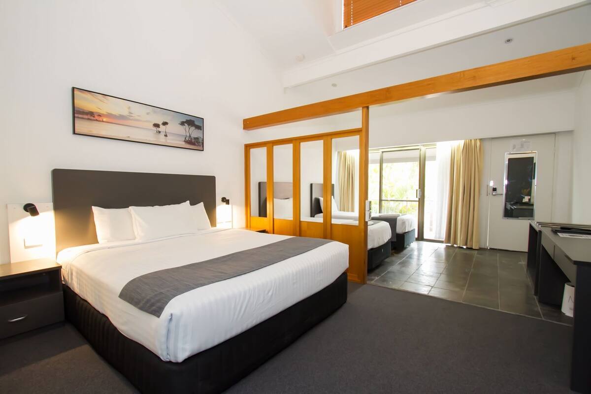 A modern hotel room with a large bed, a desk, a mirror, and a view of the outdoors.
